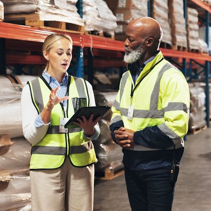 Effiziente Lagerlogistik – Teamarbeit im Warenlager Zwei Personen in Warnwesten besprechen mit einem Tablet Arbeitsprozesse in einem großen Lager.
