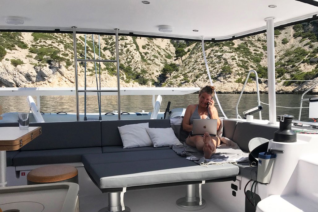 Eine Frau im gestreiften Bikini sitzt entspannt auf der gepolsterten Sitzbank eines Bootes mit ihrem Laptop und telefoniert. Im Hintergrund ist eine malerische Küstenlandschaft mit beigen Felsen und grüner Vegetation zu sehen.