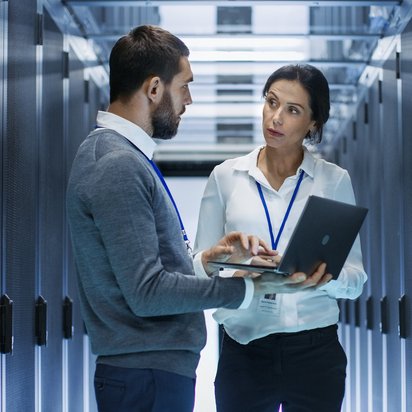 Data Analyst (TÜV) Zwei Personen stehen in einem Serverraum und besprechen technische Informationen an einem Laptop.