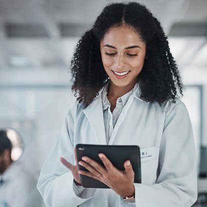 Eine Person in einem Laborkittel hält ein Tablet und steht in einer Laborumgebung.