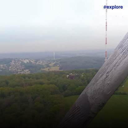 Luftaufnahme einer bewaldeten Hügellandschaft mit einer kleinen Ortschaft, aufgenommen aus großer Höhe mit einem Windradflügel im Vordergrund. Der Blick zeigt die weitläufige grüne Landschaft aus der Perspektive einer Windkraftanlage mit Funkmast im Hintergrund.