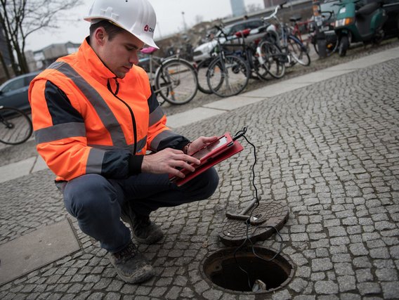 Ein Bauarbeiter in Schutzkleidung kniet auf Pflastersteinen und macht Notizen an einem geöffneten Schacht.