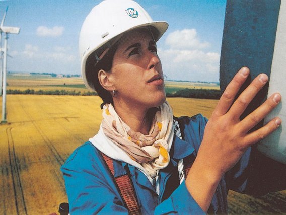 Eine Ingenieurin mit weißem Schutzhelm berührt eine Windkraftanlage vor einem Feld mit weiteren Windrädern im Hintergrund.