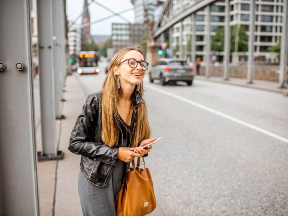 Eine lächelnde Frau mit langen blonden Haaren, schwarzer Lederjacke und Brille steht mit Smartphone und brauner Tasche an einer Bushaltestelle in einer modernen Stadtstraße.