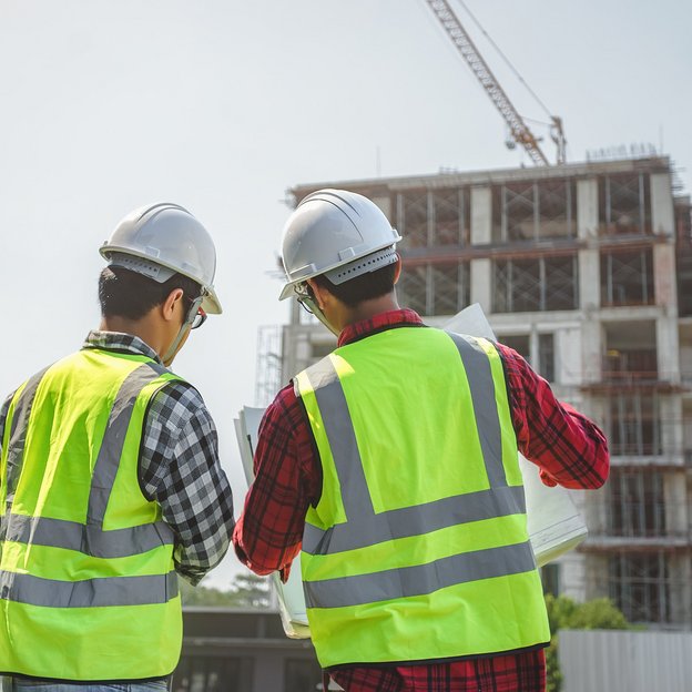 SCC - Personal: Zwei Personen des Baugewerbes befinden sich auf einer Baustelle. Beide männlichen Personen tragen Schutzkleiden wie Westen und Helme.