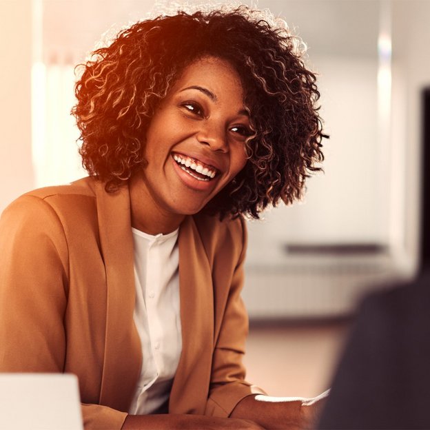 Eine Frau mit lockigem Haar und einem beigen Blazer sitzt in einem modernen Büro und spricht mit einer anderen Person. Im Vordergrund ist ein Laptop zu sehen. Das Bild vermittelt eine professionelle Coaching-Situation. 