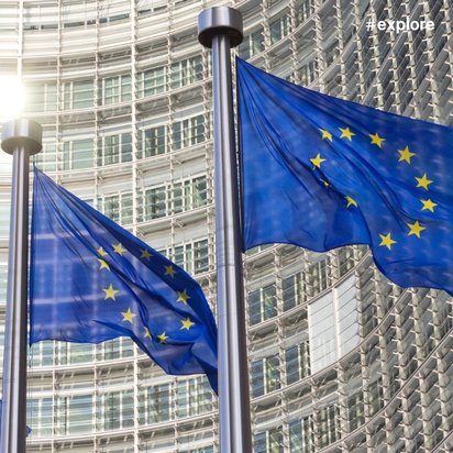 EU-Flaggen wehen vor dem Berlaymont-Gebäude der Europäischen Kommission in Brüssel. Charakteristische Glasfassade im Hintergrund von Sonnenlicht durchflutet.