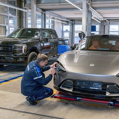 TÜV NORD Prüfer kontrolliert den Scheinwerfer eines importierten Sportwagens im Rahmen der Einzelgenehmigung, im Hintergrund ein Pick-up-Fahrzeug auf dem Prüfstand.