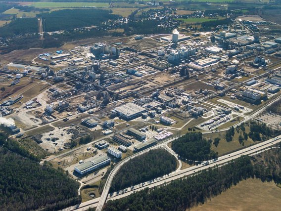 Luftaufnahme eines großen Industriekomplexes mit zahlreichen Gebäuden, Anlagen und Tanks, umgeben von Wald und Feldern sowie Verkehrswegen.