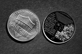 Eine US-amerikanische Ein-Cent-Münze liegt neben einer runden Platte mit kulturellen und wissenschaftlichen Informationen. Die Münze zeigt die Inschrift "E Pluribus Unum" und "One Cent". Die Platte ist mit Texten, Diagrammen und Bildern versehen und liegt auf einer strukturierten Oberfläche.