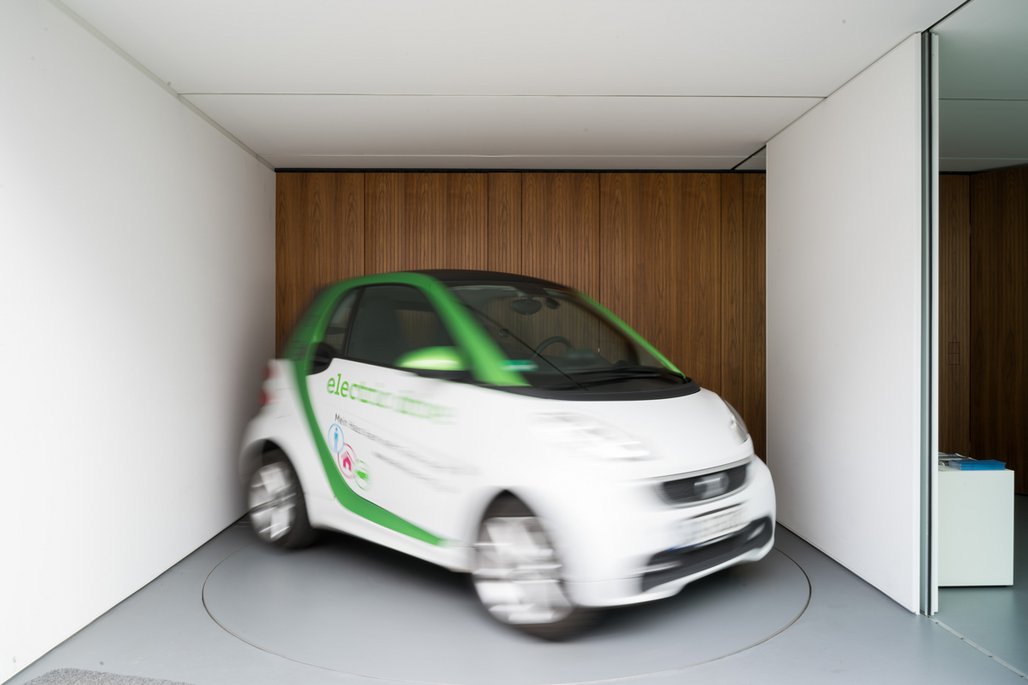 Ein kleines, weißes Elektroauto mit grüner Lackierung steht in einer kleinen Garage mit Holzverkleidung. Das Bild ist leicht verschwommen, was Bewegung andeutet.