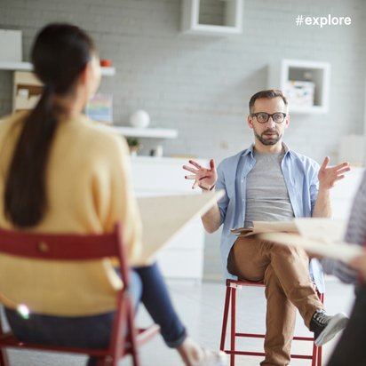 Person seated in a group discussion, speaking with hand gestures while others listen in a meeting room