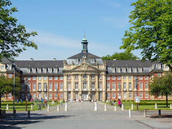 Schloss Münster mit Vorplatz und Personen bei sonnigem Wetter, eingerahmt von Bäumen