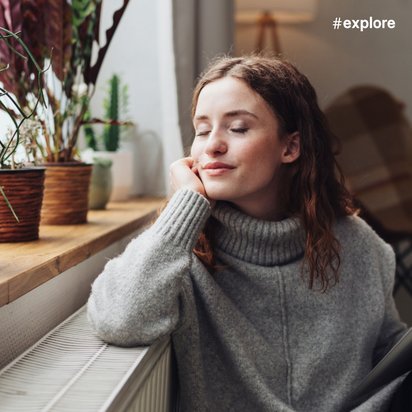 Eine junge Frau mit braunem Haar sitzt entspannt in einem grauen Rollkragenpullover an einem Fenster mit Topfpflanzen. Sie hat die Augen geschlossen, stützt den Kopf auf die Hand und hält ein Tablet.