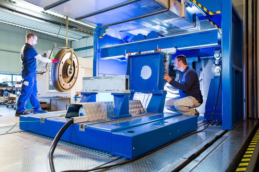 In einer industriellen Werkstatt arbeiten zwei Personen an einer großen blauen Maschine. Eine Person steht und bedient einen Kran, um ein großes, rundes Metallteil zu bewegen. Die andere Person hockt und überprüft die Positionierung des Teils in der Maschine. Die Umgebung ist gut beleuchtet und sauber, mit metallischen Oberflächen und technischen Geräten im Hintergrund.
