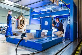 In einer industriellen Werkstatt arbeiten zwei Personen an einer großen blauen Maschine. Eine Person steht und bedient einen Kran, um ein großes, rundes Metallteil zu bewegen. Die andere Person hockt und überprüft die Positionierung des Teils in der Maschine. Die Umgebung ist gut beleuchtet und sauber, mit metallischen Oberflächen und technischen Geräten im Hintergrund.