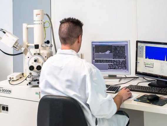 Ein Labormitarbeiter in weißem Kittel arbeitet an einem Rasterelektronenmikroskop (S-3000N) mit zwei Computermonitoren. Die Bildschirme zeigen mikroskopische Aufnahmen und Analysedaten zur Materialuntersuchung.