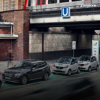 Carsharing-Fahrzeuge (BMW SUV und zwei weiße Smart-Autos) parken in grün markierten Bereichen unter einer U-Bahnbrücke.