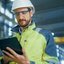 Person in Sicherheitskleidung und Schutzhelm arbeitet mit einem Tablet in einer industriellen Umgebung.