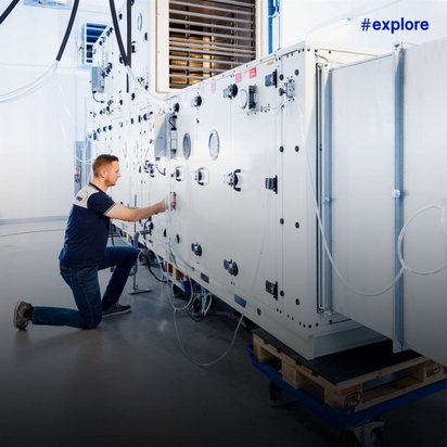 Ein kniender Techniker bedient das Kontrollpanel einer großen Klimaanlage mit Lüftungsschlitzen und zahlreichen technischen Anschlüssen.
