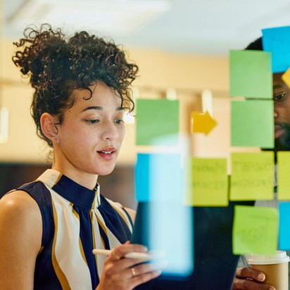 Frau arbeitet an einer Glaswand mit bunten Klebezetteln und Tablet in einem kreativen Meeting