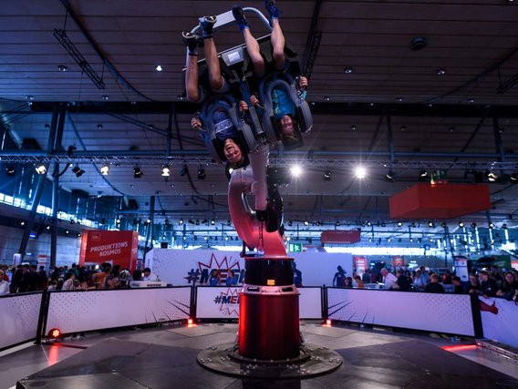 Ein spektakuläres Fahrgeschäft oder Simulator, bei dem Menschen in einer mechanischen Vorrichtung kopfüber gedreht werden. Die Installation befindet sich in der Messehalle mit vielen Zuschauern.
