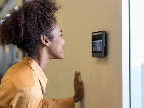 Eine Frau mit lockigem Haar in orangefarbener Bluse nutzt ein Zugangskontrollsystem. Ihr Profil ist zu sehen, während sie vor einem Wanddisplay steht.