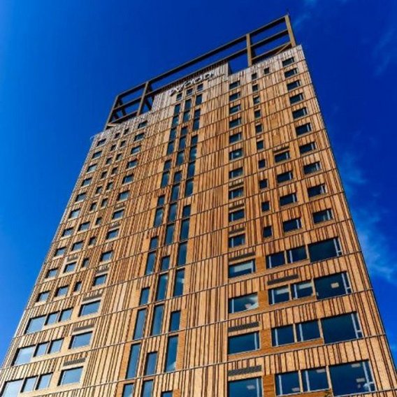 Modernes Holzhochhaus mit vertikaler Holzlamellenfassade und asymmetrisch angeordneten Fenstern vor tiefblauem Himmel, am Fuß grüne Bäume und "Wood Hotel" Schriftzug.