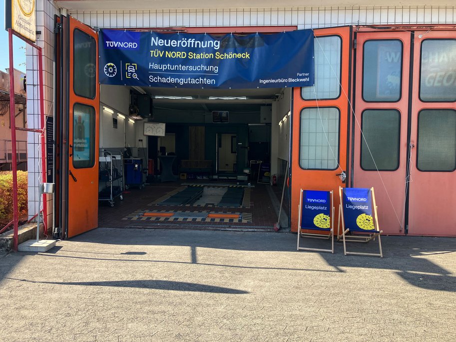 Offene Werkstatthalle mit Banner zur Neueröffnung der TÜV NORD Station Schöneck und zwei blauen Liegestühlen vor dem Eingang.