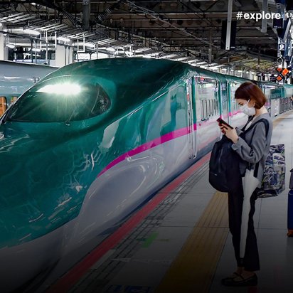 Grün-weißer Shinkansen-Hochgeschwindigkeitszug mit rosa Streifen am Bahnsteig in Japan. Wartende Fahrgäste mit Masken und Gepäck stehen vor dem stromlinienförmigen Zug in überdachter Bahnhofshalle.