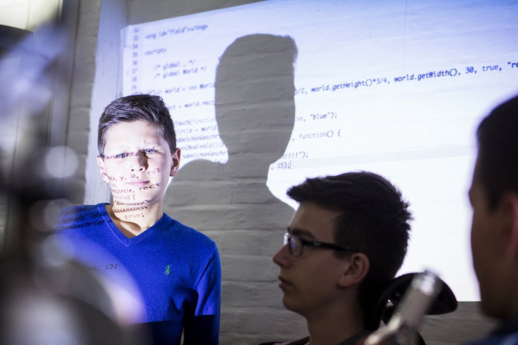 Ein Junge in blauem Pullover mit Programmcode auf sein Gesicht projiziert, daneben ein weiterer Junge mit Brille.