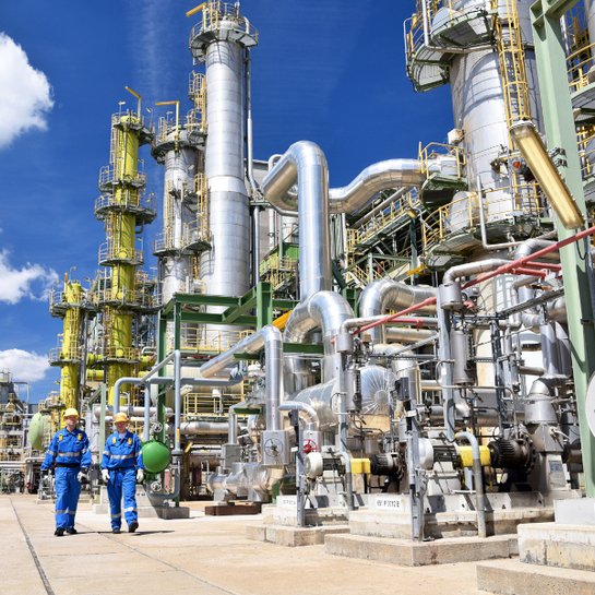 Zwei Männer in blauen Arbeitsanzügen und mit weißen Schutzhelmen laufen nebeneinander vor dem Hintergrund eines Chemieparks. Es sind diverse technische Anlagen unter blauen Himmel zu sehen.