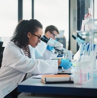 TUEV_NORD_Laborarbeit Mehrere Personen in Laborkitteln arbeiten in einem modernen Labor, eine Forscherin schaut durch ein Mikroskop.