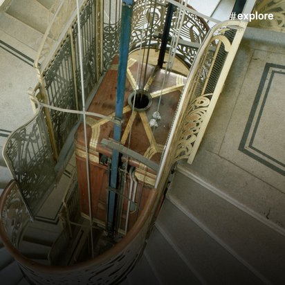 Draufsicht auf einen kunstvoll verzierten Aufzugschacht in einem eleganten Gebäude, der dekorative Schmiedeeisengitter, eine hölzerne Aufzugskabine und eine spiralförmige Treppe darum herum zeigt.