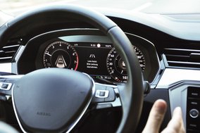 Close-up of a vehicle cockpit featuring a digital display with a lane assist warning: "Please take over steering." The steering wheel is visible in the foreground, and the dashboard displays various indicators such as speed and RPM.