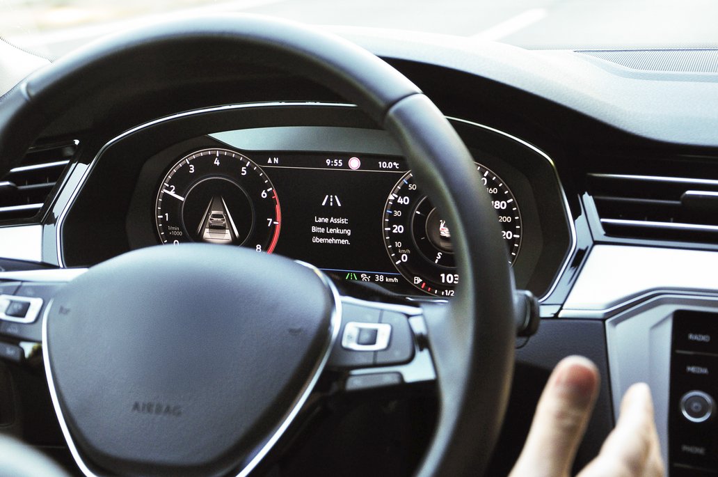 Close-up of a vehicle cockpit featuring a digital display with a lane assist warning: "Please take over steering." The steering wheel is visible in the foreground, and the dashboard displays various indicators such as speed and RPM.
