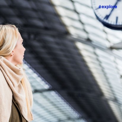 Blonde Frau in brauner Winterjacke und beigem Schal blickt nach oben in moderner Bahnhofshalle mit Glasdach und Uhr.