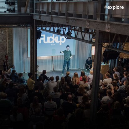 Eine Veranstaltung mit "Fuckup" im Hintergrund projiziert, bei der eine Person auf der Bühne vor einem großen Publikum steht.