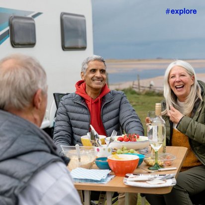Vier ältere Freunde genießen ein geselliges Essen mit Wein vor ihrem Wohnmobil bei einem malerischen Strandausblick.