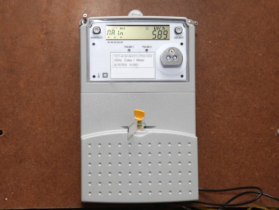 Elektrizitätszähler: Ein weißer digitaler Stromzähler mit LCD-Display, der 589 kWh anzeigt. Er hat eine graue Rückseite mit Löchern und einen gelben Drehknopf.