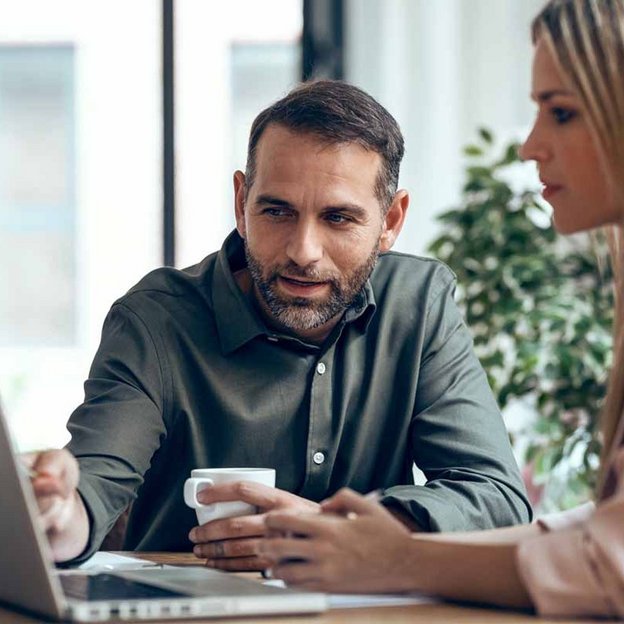 Zwei Personen diskutieren über ein Laptop an einem Tisch, während eine von ihnen eine Tasse hält. Die Szene zeigt eine konzentrierte Arbeitsatmosphäre im Rahmen finanzieller Förderung.