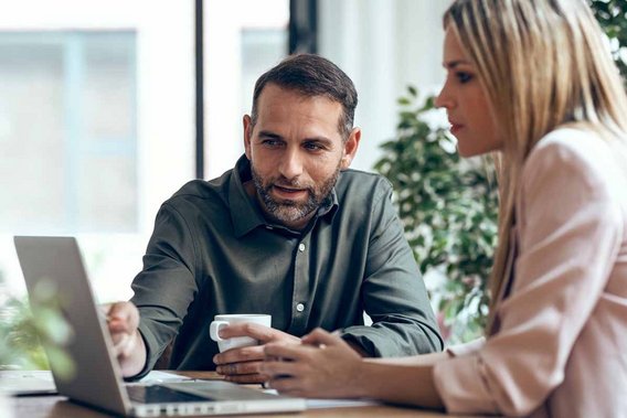 Zwei Personen diskutieren über ein Laptop an einem Tisch, während eine von ihnen eine Tasse hält. Die Szene zeigt eine konzentrierte Arbeitsatmosphäre im Rahmen finanzieller Förderung.