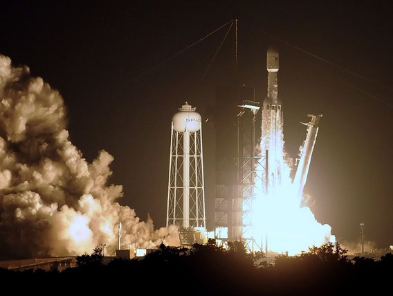 A SpaceX Falcon Heavy rocket launches at night from the launch pad, creating massive clouds of white exhaust smoke and bright flames. The rocket and launch tower are illuminated against the dark sky with the SpaceX water tower visible nearby.