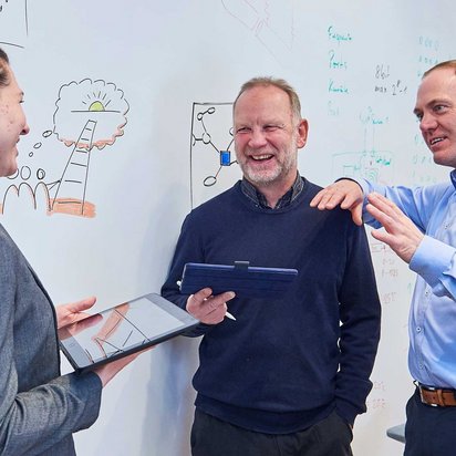 Drei Personen diskutieren in einem Büro vor einer Whiteboard-Wand. Zwei Personen halten Tablets oder digitale Geräte, während sie sich über eine gezeichnete Idee austauschen.