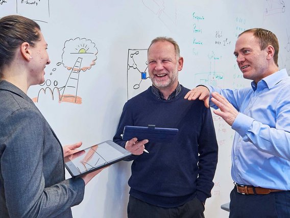 Drei Personen diskutieren in einem Büro vor einer Whiteboard-Wand. Zwei Personen halten Tablets oder digitale Geräte, während sie sich über eine gezeichnete Idee austauschen.
