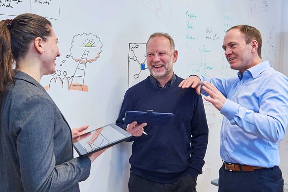 Drei Personen diskutieren in einem Büro vor einer Whiteboard-Wand. Zwei Personen halten Tablets oder digitale Geräte, während sie sich über eine gezeichnete Idee austauschen.