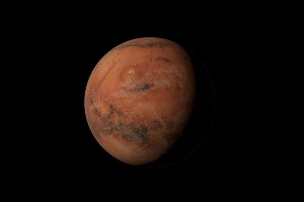 Der Planet Mars schwebt vor schwarzem Weltraum-Hintergrund und zeigt seine charakteristische rostbraune Oberfläche mit dunklen Flecken und Kratern. Die Planetenoberfläche ist teilweise von Sonnenlicht beleuchtet.