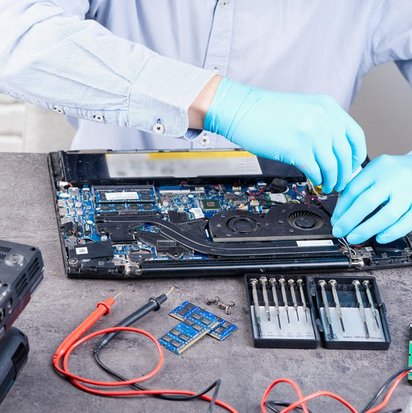 Eine Person mit blauen Handschuhen repariert einen Laptop, dessen Inneres geöffnet ist. Verschiedene Werkzeuge und Komponenten liegen auf dem Tisch.