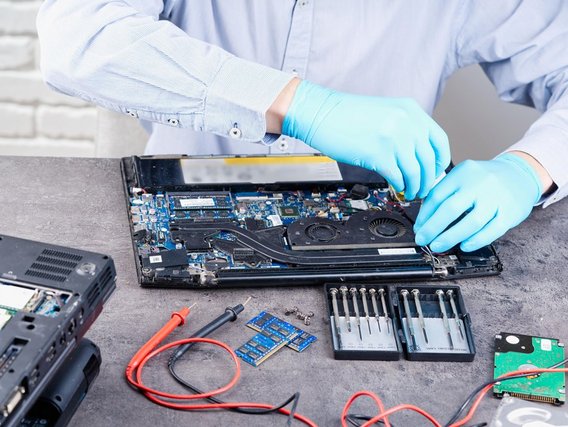 Eine Person mit blauen Handschuhen repariert einen Laptop, dessen Inneres geöffnet ist. Verschiedene Werkzeuge und Komponenten liegen auf dem Tisch.