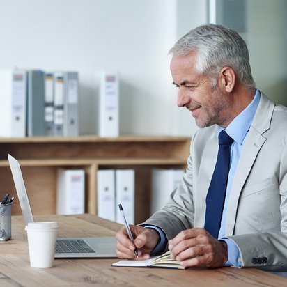 Professionelles Arbeiten im digitalen Büro. Älterer Mann im Anzug sitzt am Schreibtisch, lächelt und arbeitet mit Laptop und Notizblock.